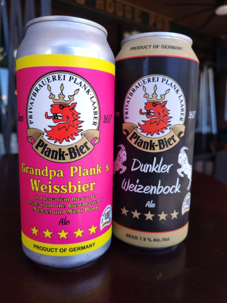 Two cans of Plank Bier's Grandpa Plank's Weissbeer and Dunkel Heizenbock Ale sit on an outdoor pub table at Bog House Pub